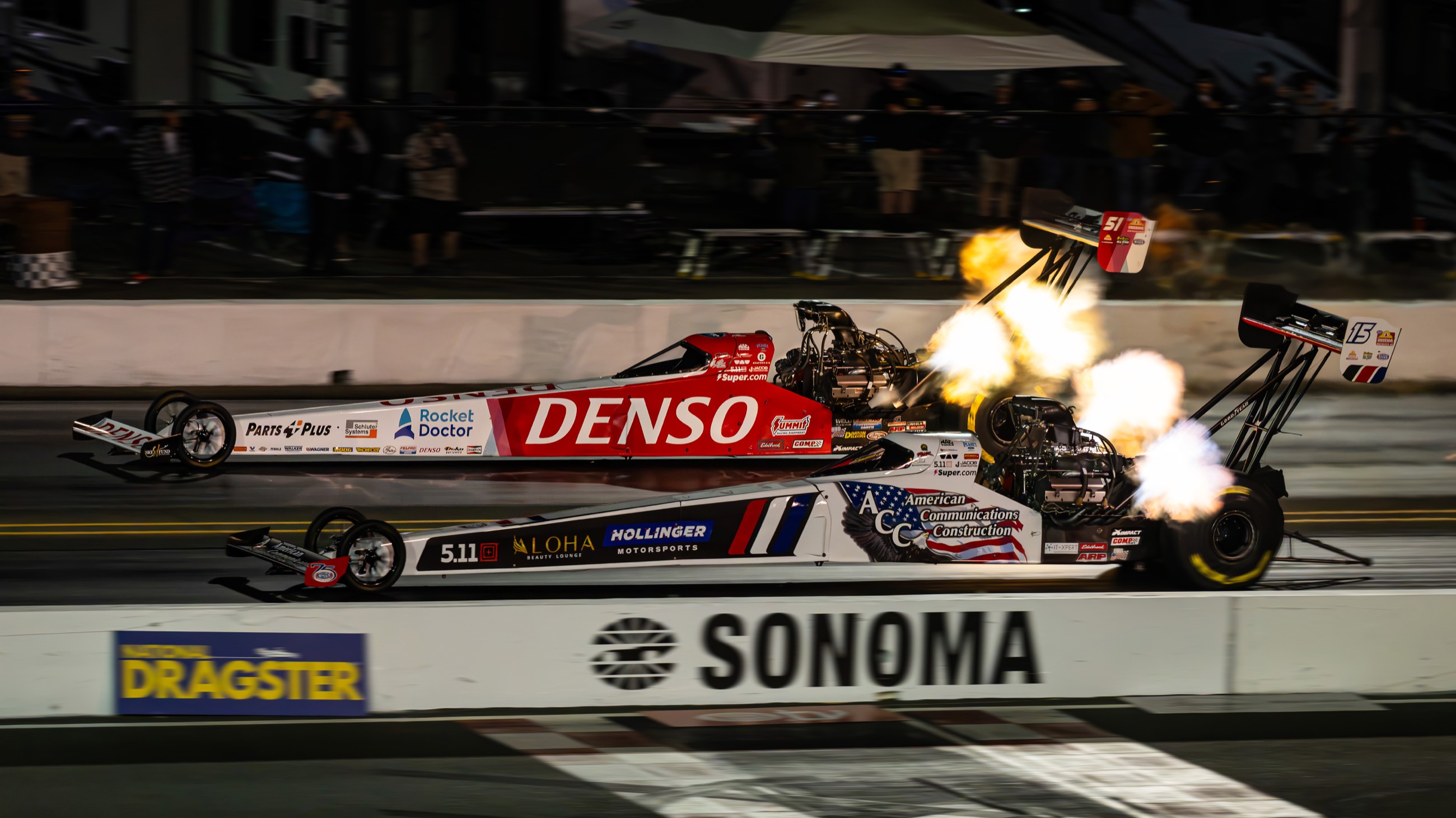 Two top fuel dragsters launching side by side at night at Sonoma Raceway, header flames lighting both lanes.