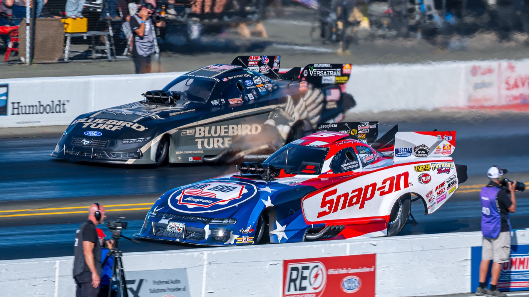 A red-white-and-blue funny car and a black funny car leaving the starting line together.
