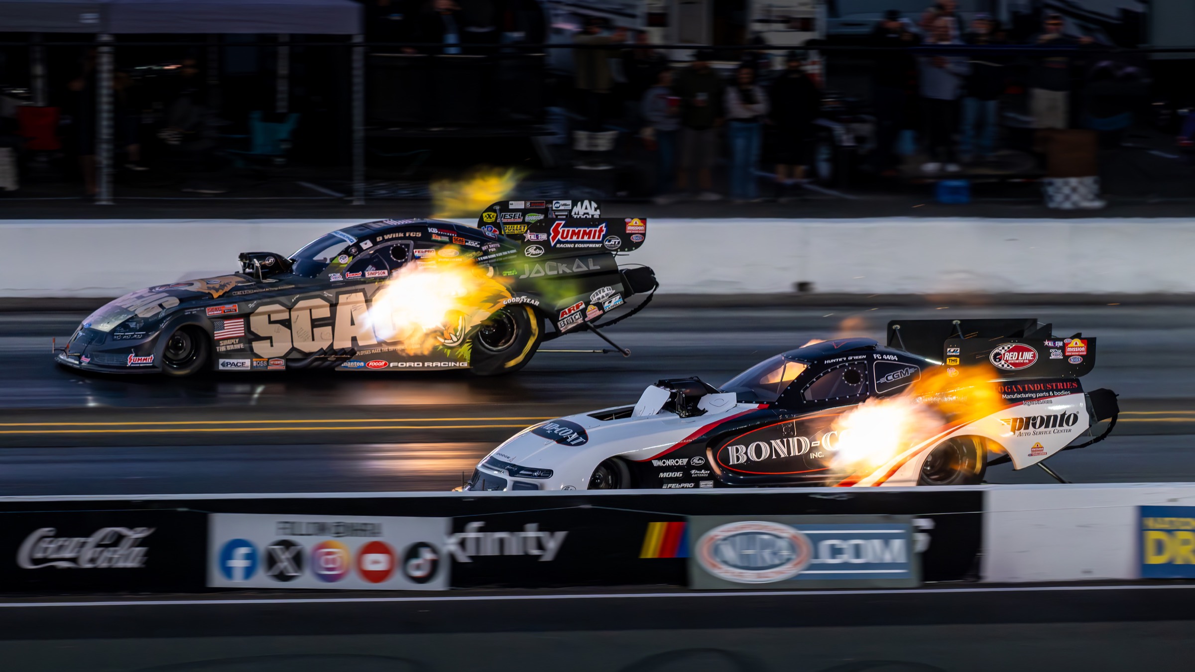 Two nitro funny cars racing side by side at dusk, exhaust headers flaming on both cars.