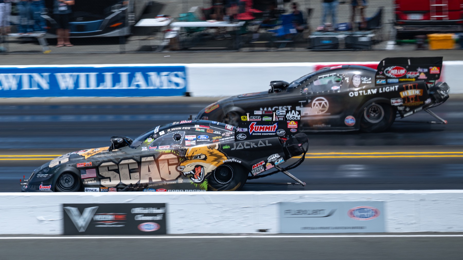 Two funny cars racing side by side past a sponsor wall.