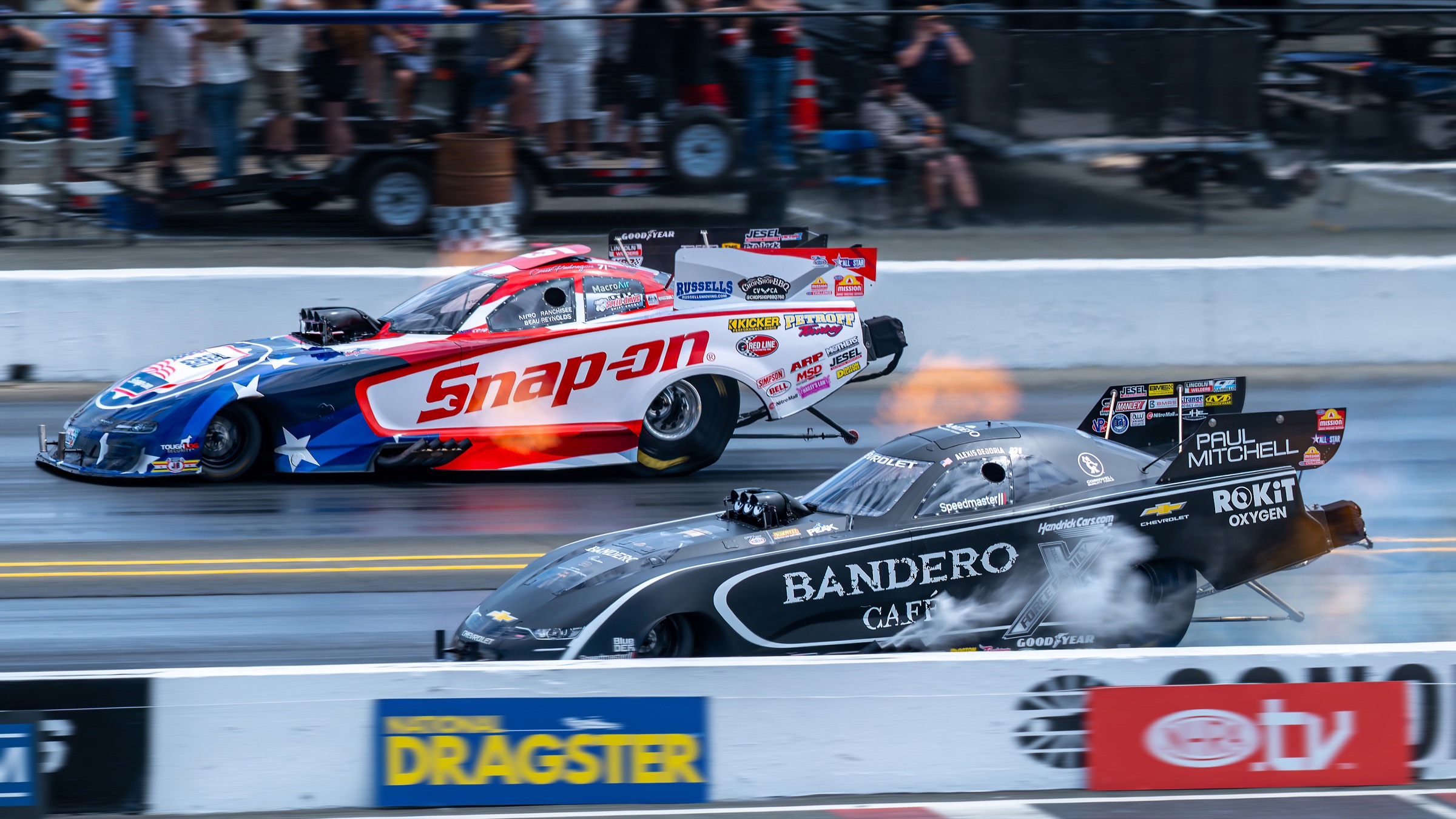 A red-white-and-blue funny car and a black funny car side by side at half track.