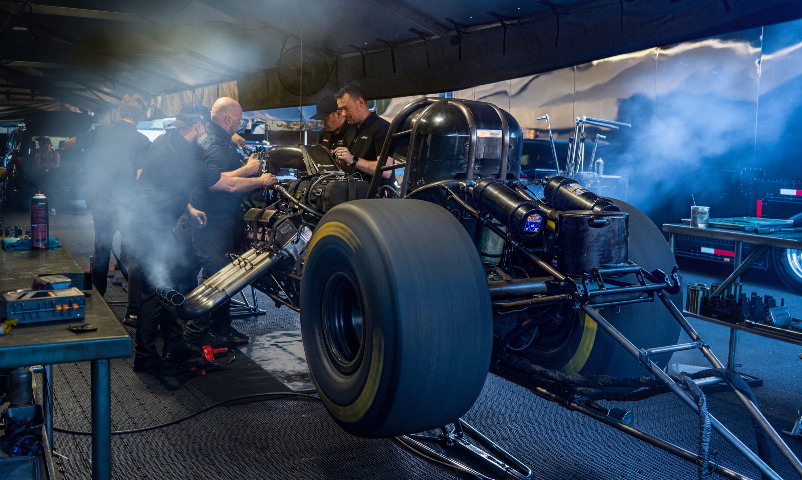 A pit crew working over an exposed top fuel engine between rounds, smoke hanging in the pit tent.